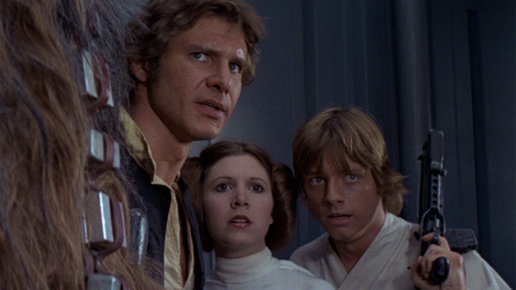 Cena de Star Wars Episódio IV: Uma Nova Esperança. Um trio de heróis se reúne numa parede na frente de uma parede metálica, observando algo além da câmera. Da esquerda para a direita, Han (Harrison Ford), Leia (Carrie Fisher) e Luke (Mark Hamill). Han é um homem caucasiano de cabelos castanhos, usando um colete preto por cima de uma camisa bege. Leia é uma mulher caucasiana de cabelos castanhos arranjados em dois coques simétricos e cheios ao seu lado, usando um vestido branco de gola alta. Luke é um homem caucasiano de cabelos loiros, usando um robe branco e segurando uma arma futurista erguida para cima com sua mão esquerda. Na frente de Han, podemos ver o peito peludo de Chewbacca, um alienígena animalesco de pelos pretos e castanhos, com uma bandoleira de munição cruzando os fios.