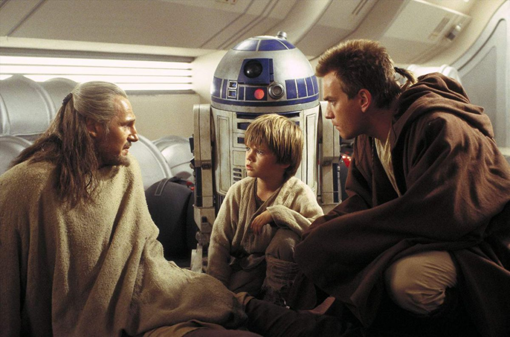 Cena de Star Wars Episódio I: A Ameaça Fantasma. Três pessoas se ajoelham no chão. Da esquerda para a direta: Qui Gon Jinn (Liam Neeson), Anakin Skywalker (Jake Lloyd) e Obi-Wan Kenobi (Ewan McGregor). Qui Gon é um homem caucasiano, de cabelos longos e loiros e uma pequena barba, usando um poncho bege grosso e calças marrons. Anakin é uma criança caucasiana de cabelos loiros, usando robes beges e grossos, com a mão esquerda apoiada em seu joelho. Obi-Wan é um homem caucasiano de cabelos castanhos, com uma fina trança do lado direito de sua cabeça. Ele usa um robe marrom grande por cima de uma roupa branca. Atrás de Anakin está R2-D2, um andróide branco e azul em forma cilíndrica com topo esférico e duas extensões dos lados para se apoias de pé. Em seu domo há uma lente preta coberta por uma placa azul e uma luz vermelha acesa embaixo. Atrás deles vemos o interior futurista de uma espaçonave, branca e angular.