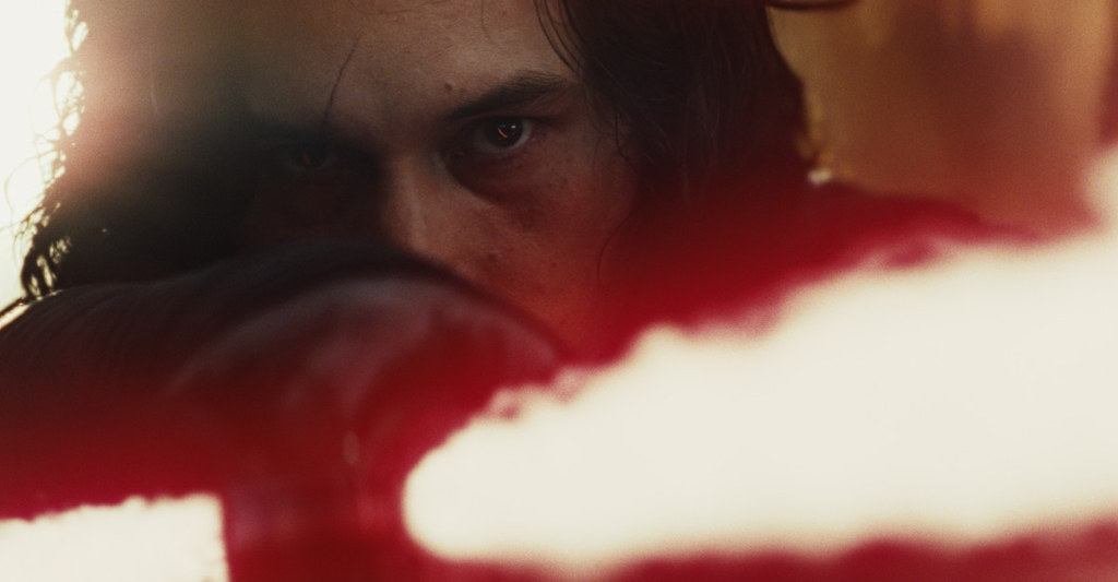 Cena de Star Wars: Os Últimos Jedi. Kylo Ren (Adam Driver) aponta seu sabre de luz na direção da câmera. Kylo é um homem caucasiano, de cabelos negros, com uma cicatriz que corta seu olho direito, olhando fixamente para frente enquanto segura seu sabre de luz com a mão direita. O sabre de Kylo é vermelho e instável, variando de volume conforme se estende para frente e para os lados, com um par de guardas também formadas por feixes de luz. Sua mão, envolta em uma luva negra, cobre a metade inferior de sua boca ao segurar o sabre apontado para frente. Atrás dele, podemos ver fora de foco um fundo amarelado, com uma luz vindo da parte esquerda da tela.