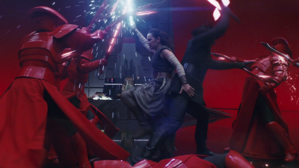 Cena de Star Wars: Os Últimos Jedi. Kylo Ren (Adam Driver) e Rey (Daisy Ridley) lutam contra a guarda pretoriana do Líder Supremos Snoke (Andy Serkis), um de costas para o outro. Rey, virada para a esquerda, é uma mulher caucasiana de cabelos negros amarrados atrás de sua cabeça, usando robes cinzentos sem mangas e tecidos brancos amarrados em seus braços. Ela segura na mão direita o cabo de um sabre de luz azul, mantida horizontalmente enquanto se defende de dois guardas, que a jogam levemente para trás. Sua mão esquerda toca a coxa de Kylo para manter o equilíbrio enquanto sua perna esquerda não toca o chão. Ela usa um botas marrons por cima de uma calça cinzenta e um cinto marrom na cintura. Kylo, virado para a direita, é um homem caucasiano de cabelos negros, usando robes inteiramente negros. Sua mão direita está erguida para cima, descendo seu sabre vermelho e instável de encontro à um dos guardas. Além da lâmina composta por um feixe de luz, duas guardas menores formam um ângulo reto na base do cabo, formando uma guarda para o sabre. Os guardas pretorianos estão vestidos com armaduras vermelhas e lisas por cima de robes vermelhos e roupas pretas. Eles empunham espadas, adagas e lanças, todas com um fio vermelho na lâmina, que produzem faíscas ao se encontrar com os sabres de luz. Vemos uma dessas lâminas na margem direita da tela, se encontrando com a armadura do guarda que enfrenta Kylo, produzindo faíscas em contato. Atrás deles, há um pano de fundo cavernoso e vermelho, partido ao meio pelo trono imenso e futurista do Líder Supremo, que jaz partido pela metade no chão, com a metade superior ainda sentada no trono.