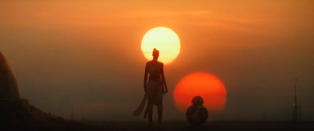 Cena de Star Wars: A Ascensão Skywalker. Rey (Daisy Ridley) e o dróide BB-8 observam o nascer de dois sóis no planeta desértico de Tatooine. Rey, à esquerda, é uma mulher caucasiana, com cabelos pretos presos em um coque, com um dos sóis, amarelo, destacando o topo de sua cabeça na distância. Ela usa vestes brancas que balançam com o vento, um cinto escuro com o cabo de um sabre de luz na direita. BB-8, à direita, tem o corpo no formato de uma esfera com um domo no topo, é destacado pelo segundo sol, maior e mais baixo que o de Rey, alaranjado. BB-8 chega a altura dos joelhos de Rey. Na direita, podemos ver um tipo de torre na distância com algum tipo de aparato no topo, enquanto na direita, uma pequena elevação no solo dá indícios de algum tipo de construção. Fora essas coisas, a paisagem é completamente plana