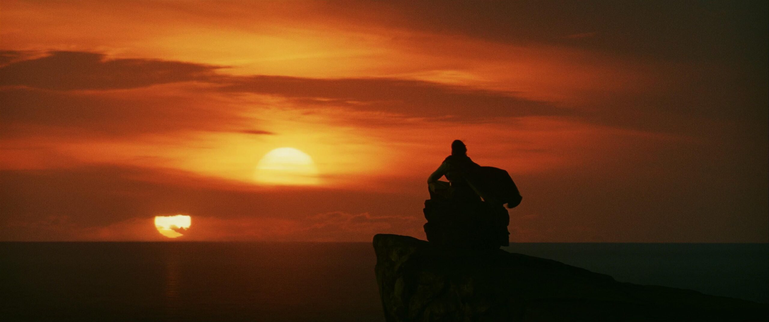 Cena de Star Wars: Os Últimos Jedi. Luke (Mark Hamil) está sentado de pernas cruzadas em cima de uma pedra que repousa em um rochedo que encara um mar tranquilo. Ele ocupa a parte direita da imagem, de costas para a tela, e observa ao longe dois sóis se pondo na distância