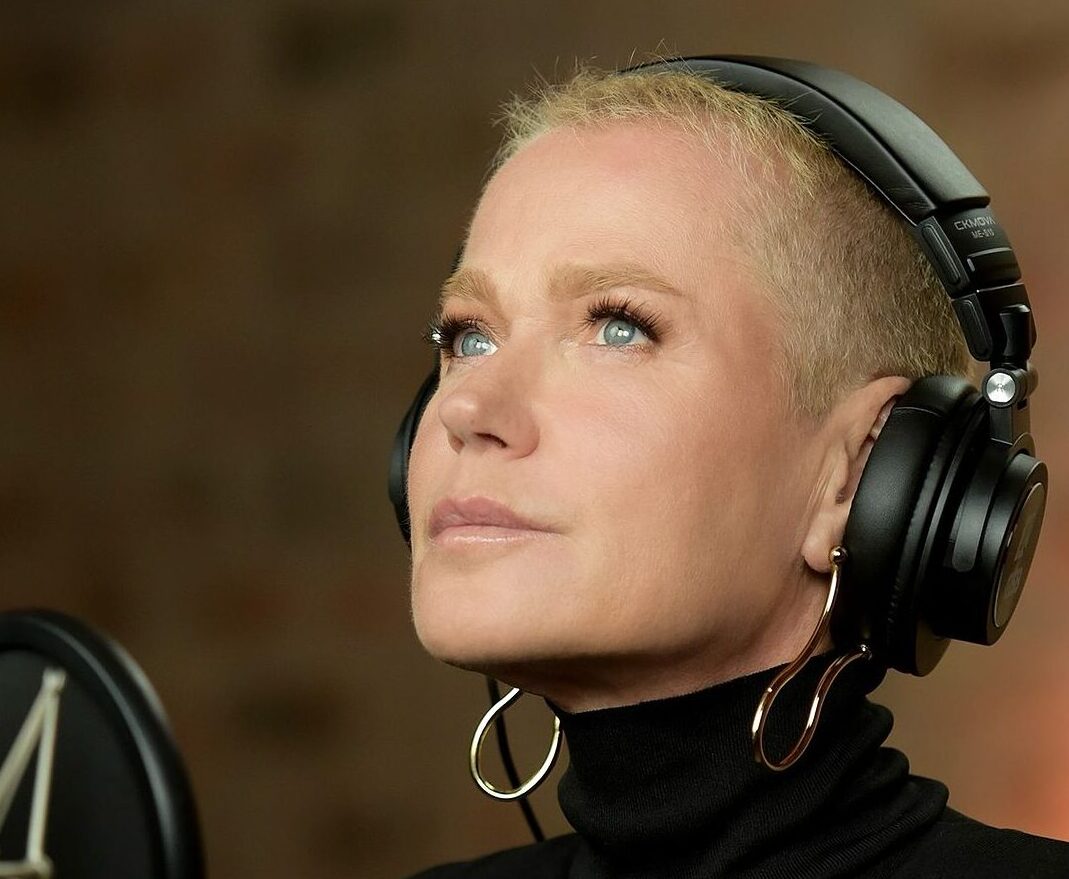 A imagem mostra o rosto de perfil da apresentadora Xuxa Meneghel, uma mulher branca, de cabelos loiros raspados e olhos azuis. Ela veste uma blusa preta de gola alta, que cobre o pescoço, e usa brincos de prata com design torcido. À sua frente, há um microfone, e ela está com fones de ouvido, sugerindo que a cena ocorre em um estúdio de gravação.