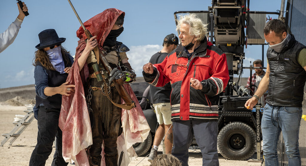 Foto dos bastidores do filme Furiosa: Uma Saga Mad Max. Anya Taylor-Joy e George Miller discutem no deserto enquanto profissionais ao seu redor preparam a cena. Anya, à esquerda, é uma mulher caucasiana, vestida como a personagem Furiosa, usando uma capa vermelha desbotada sobre seu corpo e cobrindo sua cabeça. Ela usa uma camiseta branca suja por baixo de um macacão de couro, segurando uma espingarda com as duas mãos, apontada para cima. Em sua cintura há um cinto adornado pelo emblema de uma caveira flamejante e uma máscara preta cobre sua boca. Seu braço esquerdo é adornado por um aparelho mecânico e faixas verde claras. George, à direita, é um homem caucasiano idoso, usando uma jaqueta vermelha com detalhes pretos, usando uma calça azul e gesticulando para Anya com suas duas mãos. À sua direita, um comem de colete preto e máscara branca está olhando para algo no chão. À esquerda de Anya, uma profissional está reposicionando sua capa vermelha. Ela usa um chapéu preto e usa uma máscara azul clara e óculos pretos, vestida com uma camisa azul e calça preta. Atrás deles, vemos dois homens trabalhando em um equipamento em cima de um carrinho preto. Ao fundo, um céu azul marcado por poucas nuvens se estende, quebrado apenas por uma pequena colina no horizonte.