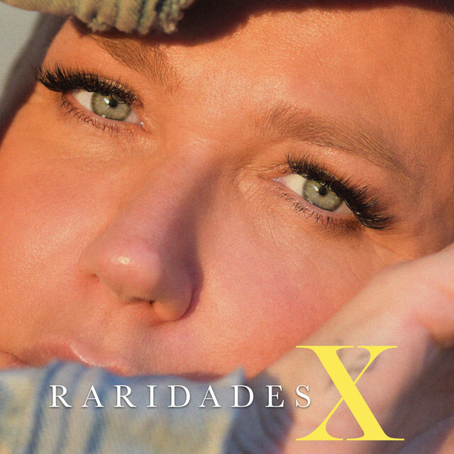 A imagem exibe a capa do novo álbum de Xuxa, com uma foto muito próxima do rosto da artista. Xuxa entrelaça o rosto com os braços, destacando uma pose que enfatiza os cílios postiços e os olhos azuis, que encaram diretamente o observador. O título "Raridades" aparece em branco no canto inferior central, enquanto o "X" em amarelo, de tamanho maior, se destaca à direita.
