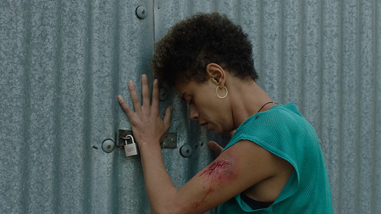 Cena do filme Continente. A imagem mostra, ao centro, uma mulher negra, de cabelos curtos escuros, aparentando cerca de 30 anos, vestindo uma camiseta azul apoiada em uma porta de metal com um cadeado na fechadura. O braço esquerdo da mulher está sangrando.