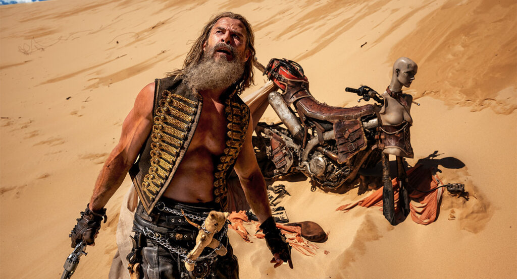 Cena do filme Furiosa: Uma Saga Mad Max: Dementus (Chris Hemsworth) está na frente de sua motocicleta, olhando confuso para algo acima das dunas. Dementus é um homem caucasiano de cabelos longos e barba grisalha. ele usa um colete preto com detalhes dourados sem camisa por baixo e calça preta. Em sua cintura, um ursinho de pelúcia bege está preso por correntes na frente de sua pelve. Na mão direita, ele segura um revólver. Atrás dele, na direita, está sua motocicleta improvisada: no lugar da frente há o busto de um manequim feminino cinza, e atrás, um tanque de combustível vermelho amarrado com faixas de couro e fivelas. A roda da frente da moto está faltando.