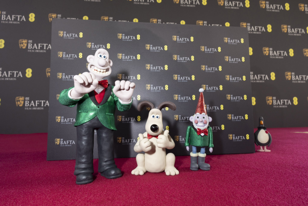 Foto dos modelos de Wallace, Gromit e Norbot no tapete vermelho do BAFTA 2025. Wallace é um homem caucasiano careca, usando um terno paletó verde por cima de um colete vermelho e uma camisa branca, com uma gravata borboleta vermelha e calças pretas. Ele está com as mãos erguidas em animação. Gromit é um beagle de pelo claro e orelhas escuras, sentado nas patas traseiras e dando jóia com as mãos. Norbot é um robô em forma de gnomo de jardim, usando um paletó verde e gravata borboleta vermelha com uma calça azul clara e botas escuras. Ele usa um chapéu cônico grande e vermelho e possui uma barba branca e bochechas vermelhas. Atrás deles, saindo de trás da parede com o logo da premiação, está o pinguim Feathers McGraw, usando uma gravata borboleta preta e uma rosa no peito.