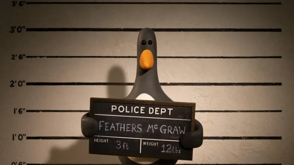 Cena da animação Wallace e Gromit: Avengança. O pinguim Feathers McGraw está tirando uma foto na frente de um fundo branco com linhas pretas horizontais marcando a altura em pés. Ele segura uma placa preta com seu nome, sua altura e seu peso, encabeçados pelo nome "Departamento de Polícia".