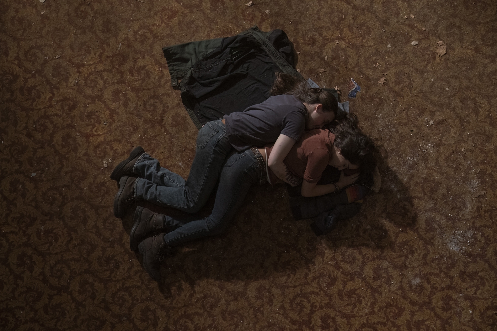 Cena da segunda temporada de The Last of Us. Ellie (Bella Ramsey) e Dina (Isabela Merced) estão deitadas no chão, abraçadas uma na outra, por cima de seus casacos. Ellie é uma mulher caucasiana jovem de cabelos negros, usando uma camiseta escura, calças jeans e botas marrons. Dina é uma mulher hispânica jovem de cabelos negros, usando uma camiseta alaranjada, calças jeans e botas marrons. Ellie, à esquerda, abraça Dina pelas costas. O chão ao redor delas é um carpete vermelho velho com decorações florais amareladas e rodeado por detritos.