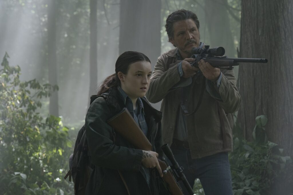 Cena da segunda temporada de The Last of Us. Ellie (Bella Ramsey) e Joel (Pedro Pascal) andam por uma floresta armados. Ellie (à esquerda) é uma mulher caucasiana jovem de cabelos pretos e longos, amarrados num rabo de cavalo. Em sua sobrancelha direita há uma pequena cicatriz perpendicular. Ela usa uma jaqueta escura por cima de uma camisa azul quadriculada e segura um rifle de caça nas mãos, apontado para baixo. Na sua mão direita, vemos a ponta de uma tatuagem escura de folha em seu pulso. Nas costas, ela carrega uma mochila. Joel (à direita) é um homem latino de meia idade, com cabelos pretos, curtos e grisalhos e uma barba rala, usando uma jaqueta bege por cima de uma camisa azul clara e uma calça jeans. Ele aponta seu rifle para a direita, com a coronha da arma apoiada em seu ombro direito. Atrás deles, vemos silhuetas de árvores iluminadas pelo Sol e, ao seu lado, arbustos iluminados. Na direita, ao lado de Joel, vemos o tronco descascado de uma árvore.