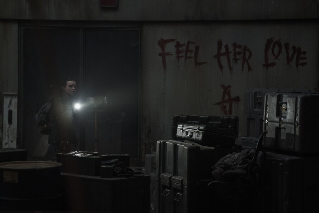 Cena da segunda temporada de The Last of Us. Ellie (Bella Ramsey) está apontando sua lanterna para frente, ofuscando parte de seu corpo. Ellie é uma mulher caucasiana jovem, de cabelos negros, usando um casaco bege e carregando uma mochila nas costas. Na sua frente podemos ver caixotes escuros iluminados e alguns barris. Atrás dela há uma porta de vidro e, na parede clara ao seu lado, uma mensagem escrita com sangue que mostra as palavras "sinta o amor dela" em inglês, junto com um símbolo oval cortado horizontalmente no meio.