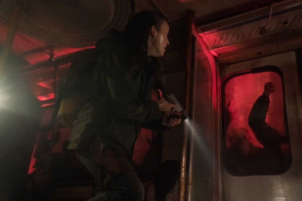 Cena da segunda temporada de The Last of Us. Ellie (Bella Ramsey) caminha dentro do vagão de um trem destruído enquanto infectados socam as portas tentando entrar. Ellie é uma mulher caucasiana jovem de cabelos negros, usando uma jaqueta bege e carregando uma mochila nas costas. Ela carrega uma lanterna na mão esquerda e uma pistola na direita, observando cautelosamente os infectados que tentam invadir o trem. Uma luz vermelha ilumina o interior, vinda de um sinalizador fora do vagão.