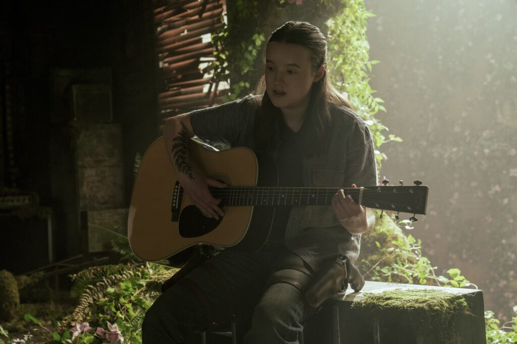 Cena da segunda temporada de The Last of Us. Ellie (Bella Ramsey) está sentada, com um violão no colo, tocando uma música em frente à uma abertura gigante na parede. Ellie é uma mulher caucasiana jovem de cabelos negros, usando uma camisa cinzenta sem mangas por cima de uma camiseta escura. Ela usa uma calça jeans e, por cima dela, na perna esquerda, vemos um coldre amarrado em sua coxa, contendo um revólver. Ela se senta em uma caixa de metal coberta por musgo virada para o lado e, atrás dela, vemos vinhas e folhas se espalhando pela abertura na parede. Uma tatuagem de folhas cobre seu antebraço direito.