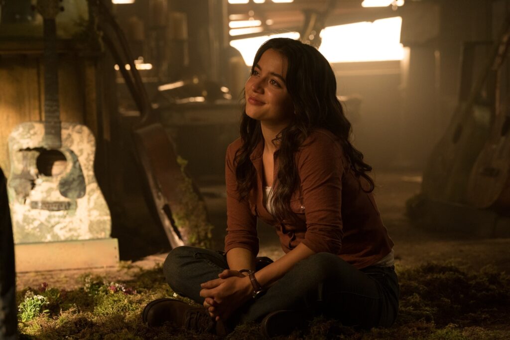 Cena da segunda temporada de The Last of Us. Dina (Isabela Merced) está sentada no chão, olhando apaixonadamente para Ellie. Dina é uma mulher hispânica jovem, de cabelos negros, usando uma camisa alaranjada por cima de uma camiseta branca. Ela está sentada na grama que cresce no chão e atrás dela podemos ver uma loja de música dilapidada, com violões arruinados de pé e uma janela aberta.