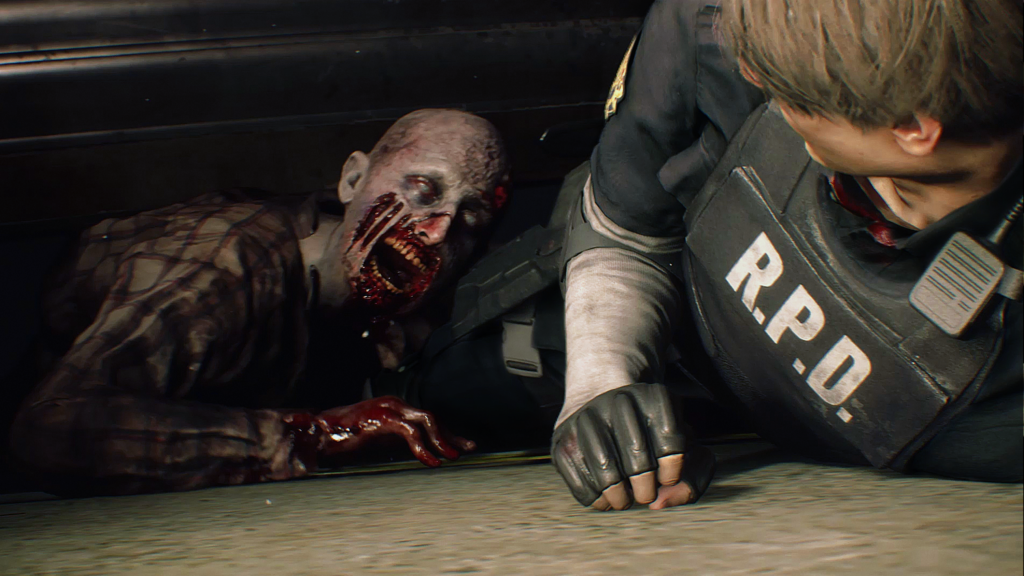 Cena do jogo Resident Evil 2: Leon está no chão, tentando escapar de um zumbi agarrado em seu pé. Leon é um homem caucasiano jovem, de cabelos loiros, usando um uniforme de polícia de Raccoon City, com a sigla R.P.D. em letras brancas no colete em seu peito. Suas mangas são cinzentas e ele usa luvas negras e braçadeiras pretas. Na parte superior esquerda de seu peito, vemos um rádio preso ao colete. O zumbi, agarrado em suas calças pretas, é careca e tem a pele enrugada e pálida, um olhar esbranquiçado e parte dos músculos da bochecha faltando, expondo a carne de sua mandíbula, ele usa uma camisa xadrez manchada e vemos sua mão direita manchada de sangue no chão. Ele está embaixo de uma grade metálica quase fechada.