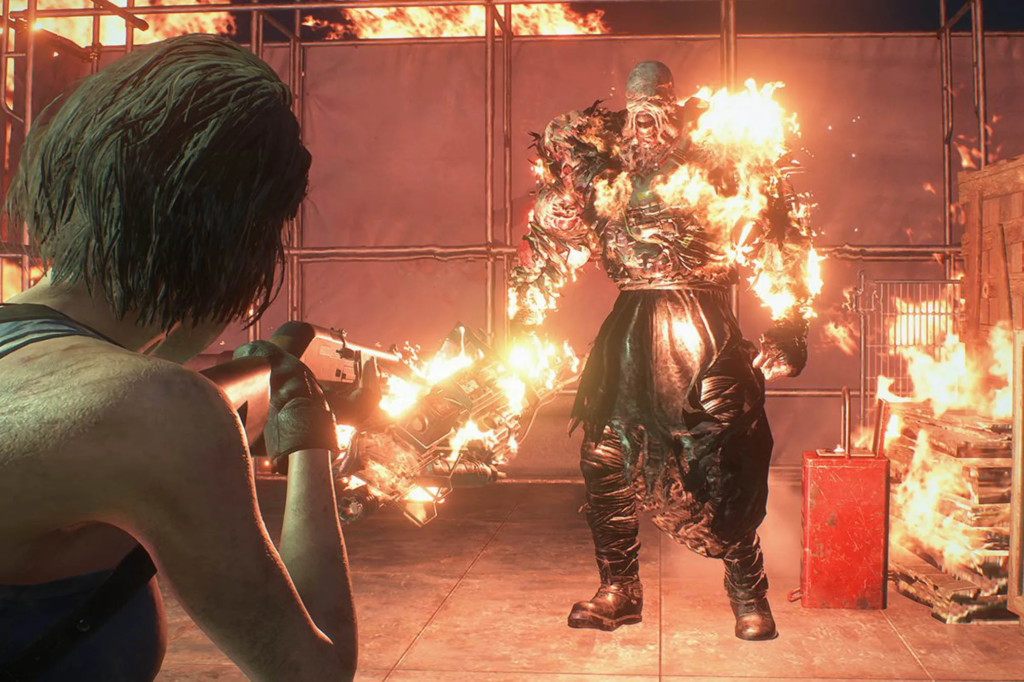 Cena do jogo Resident Evil 3: Jill Valentine mira na criatura Nemesis com sua espingarda. Jill é uma mulher caucasiana de cabelos castanhos, usando uma camiseta regata azul, um coldre preto nos ombros e luvas pretas. Ela, à esquerda da tela, segura a arma com as duas mãos, mirando no monstro na sua frente. Nemesis é um ser humanóide, com o corpo gigante revestido por um tecido preto coberto por tubos e amarrado em volta dos seus membros. Partes rasgadas desse tecido expõem partes do seu corpo desfigurado, coberto por chamas. Ele está com a boca arreganhada, esticando a pele de sua face e deixando aparecer os músculos de sua mandíbula. Na mão direita, ele segura uma grande arma metálica. À sua esquerda, vemos um galão vermelho ao lado de alguns caixote de madeira empilhados. Atrás dele, vemos um tecido azul e algumas treliças de ferro expostas, com chamas subindo atrás delas contra ao céu noturno.