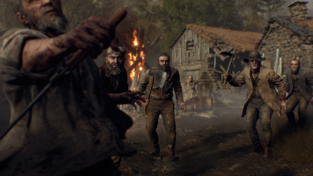 Cena do jogo Resident Evil 4: um grupo de habitantes infectados de um vilarejo se aproxima para atacar, com armas em punho. Os habitantes se vestem com roupas campestres, camisas xadrez, casacos leves, chapéus de aba e boinas, empunhando machados, facas, foices e ancinhos. Atrás deles, vemos um celeiro de madeira e pedra com a parte direita demolida e uma casa de pedra com tábuas de madeira empilhadas ao lado da parede. Entre dois habitantes, uma fogueira gigante está acesa, adornada com uma forma cruciforme com um X cruzando o eixo vertical. Atrás das casas vemos troncos de árvores amontoadas, iluminadas pela luz do Sol. O chão do vilarejo é de terra marrom.