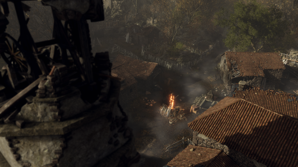 Cena do jogo Resident Evil 4: um vilarejo rural, em estado dilapidado, com uma grande fogueira queimando na parte central, em frente à uma igreja. As casas ao redor são feitas de pedra, com tetos de telha, com cercas cobrindo parte do terreno. O chão do vilarejo é de terra batida e, ao lado da fogueira, vemos um poço de água decorado com uma estela. Fora de foco, cobrindo a parte esquerda da tela, vemos uma torre de sino arruinada, com o sino faltando e a parte de cima em pedaços. Não há ninguém no vilarejo e a cena toda é iluminada pela luz do Sol.