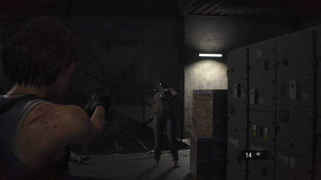 Cena do jogo Resident Evil 3: Jill Valentine dispara contra a cabeça de um zumbi dentro de um edifício. Jill é uma mulher caucasiana de cabelos castanhos, usando uma camiseta regata azul e luvas pretas, segurando uma pistola escura com as duas mãos. Na sua frente, a cabeça do zumbi é jogada para trás com o impacto da bala, mas ele permanece de pé. Sua pele é cinzenta e ele usa uma camiseta clara por cima de calças jeans, com cabelos pretos e longos. Do lado esquerdo de Jill, uma fileira de equipamentos eletrônicos cinzentos está apagada, com algumas caixas de papelão em sua frente. Atrás do zumbi, uma escada de metal escura contra uma parede de concreto, iluminada por uma lâmpada branca horizontal fixada na parede.