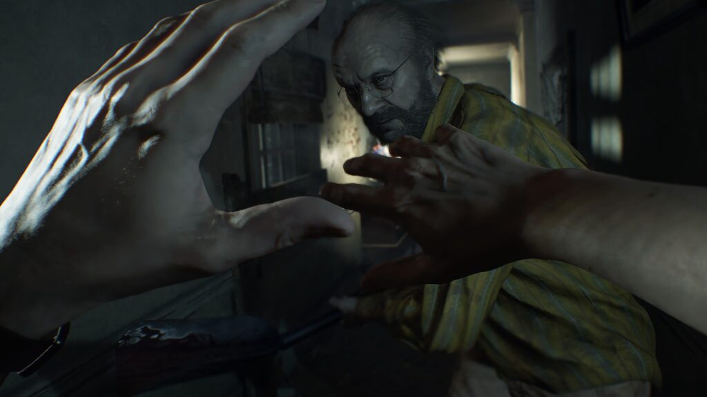Cena do jogo Resident Evil 7: biohazard. Perspectiva em primeira pessoa: Ethan ergue os braços para se proteger do ataque vindo de Jack. Ethan é um homem caucasiano e conseguimos ver suas mãos nuas erguidas em um gesto de defesa. Jack é um homem caucasiano de meia idade, com cabelo começando a rarear e barba espessa e grisalha, usando óculos e uma camisa amarela e listrada. Ele ataca com uma pá de ferro, impelindo-a contra Ethan. Atrás deles vemos o corredor de uma casa com papel de parede florido e uma janela coberta por tábuas, deixando apenas parte da luz entrar. Atrás de Jack, uma luz parece vir de um abajur.