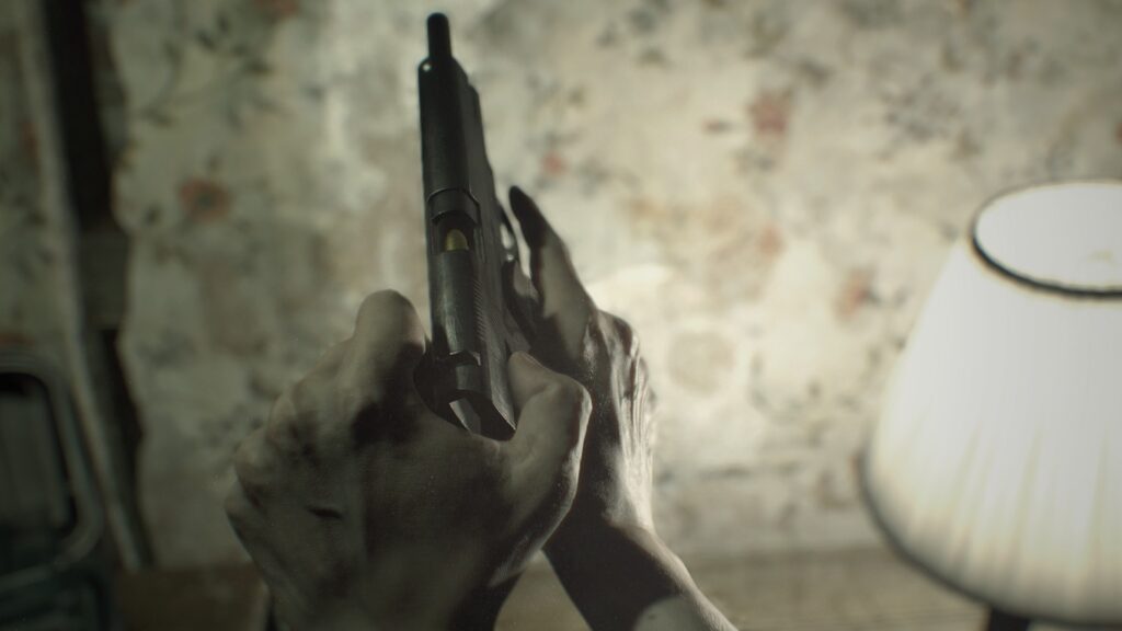 Cena do jogo Resident Evil 7: biohazard. Perspectiva em primeira pessoa: Ethan puxa o cano de uma pistola para trás para checar a bala inserida na câmara. Ethan é um homem caucasiano e vemos apenas suas mãos nuas segurando a arma. Na frente dele, um papel de parede decorado com flores fora de foco, iluminado pela luz branca de um abajur à direita.