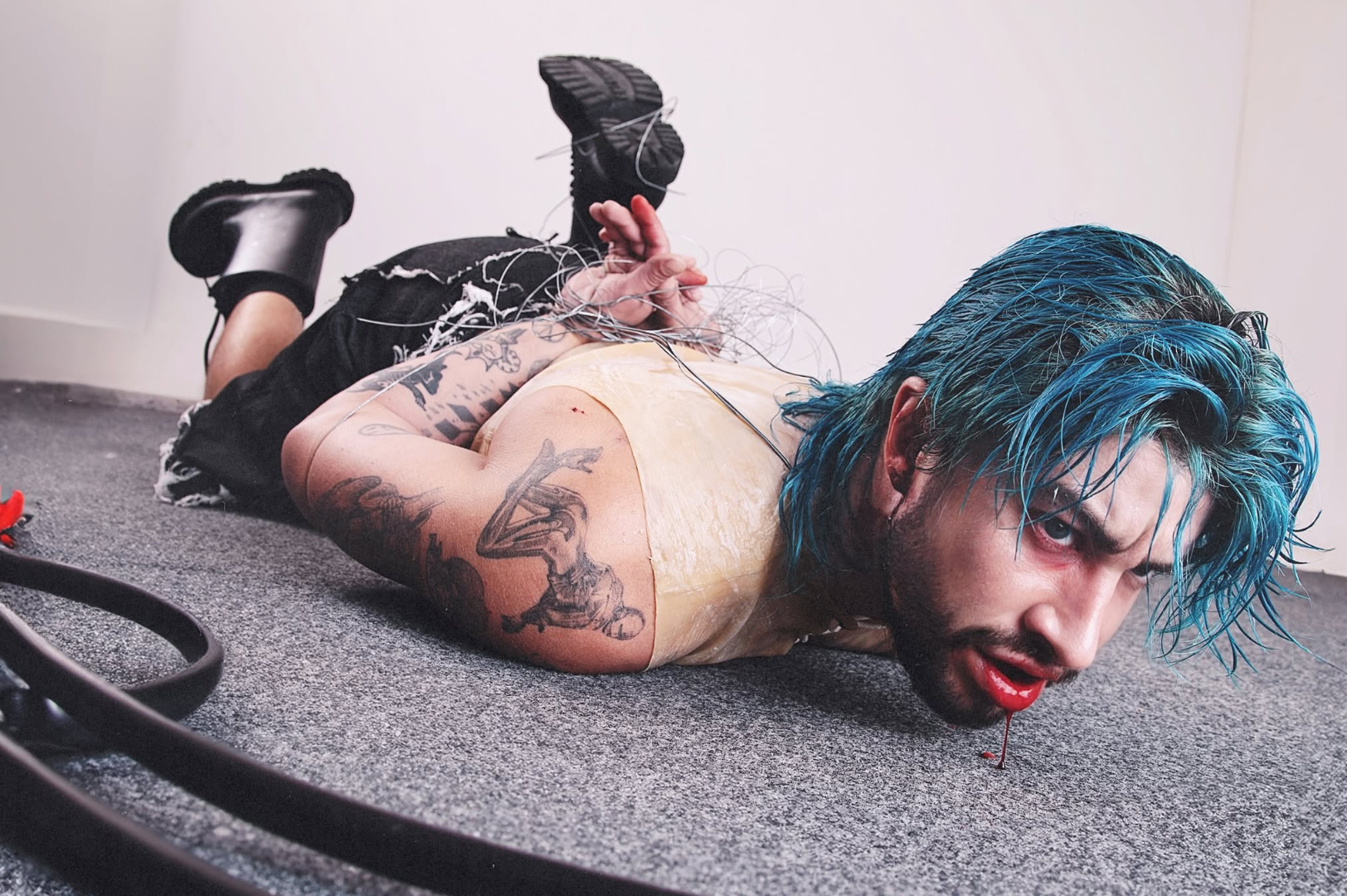 A imagem mostra o cantor CHAMELEO, um homem branco, caído no chão, usando uma peruca azul úmida. Seus braços estão amarrados, há sangue saindo de sua boca e ele exibe algumas tatuagens.