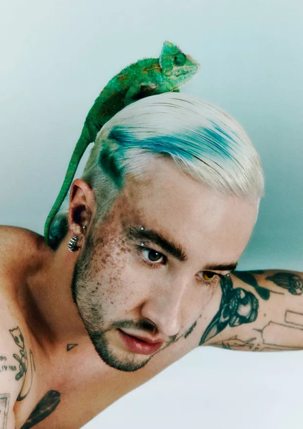 A imagem mostra um close do rosto de CHAMELEO, com um camaleão verde posicionado sobre sua cabeça. É possível observar algumas sardas no rosto do cantor, suas tatuagens no braço e acessórios prateados, como brincos e piercings na sobrancelha.