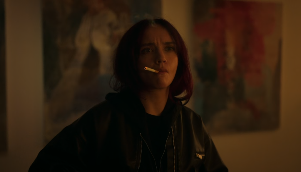 Cena da primeira temporada de The Girlfriend. Cherry (Olivia Cooke) está fumando um cigarro, olhando intensamente para um ponto além da câmera, à direita da tela. Cherry é uma mulher caucasiana de cabelos ruivos e longos, puxados para trás, usando uma jaqueta preta de couro por cima de um moletom escuro. Em sua boca vemos um cigarro aceso, apontado para cima e, atrás dela, obras de arte penduradas numa parede branca, mal iluminadas e fora de foco.