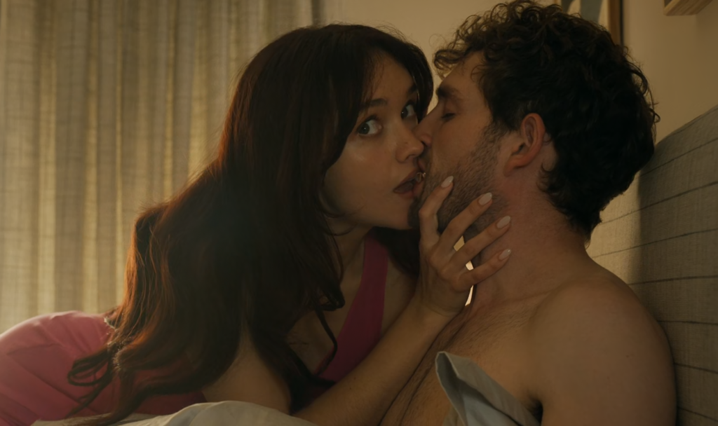 Cena da primeira temporada de The Girlfriend. Cherry (Olivia Cooke) se debruça para beijar Daniel (Laurie Davidson) enquanto desvia o olhar para algo além da tela. Cherry é uma mulher caucasiana de cabelos ruivos e soltos, usando uma camisola rosa. Daniel é um homem caucasiano com cabelos pretos e barba curta, sem camisa. Atrás deles, vemos uma cortina translúcida bloqueando parte da luz do Sol e, atrás de Daniel, a cabeceira listrada da cama onde ele está deitado.