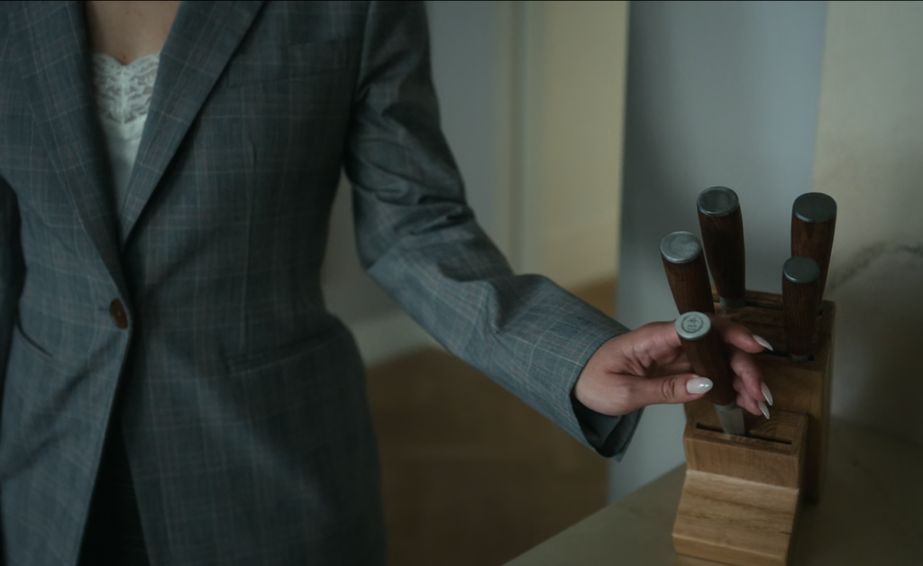 Cena da primeira temporada de The Girlfriend. Uma mão se estende e puxa uma faca de um faqueiro de madeira. A mão pertence à um corpo caucasiano e feminino, usando um paletó cinza fechado por cima de uma camiseta de renda branca. A mão possuí unhas longas, pintadas com uma cor clara e puxa delicadamente uma das cinco facas que estão no faqueiro, em cima de uma mesa de mármore. Atrás dela, vemos corredores pintados com uma cor clara e um chão de madeira polida, fora de foco.