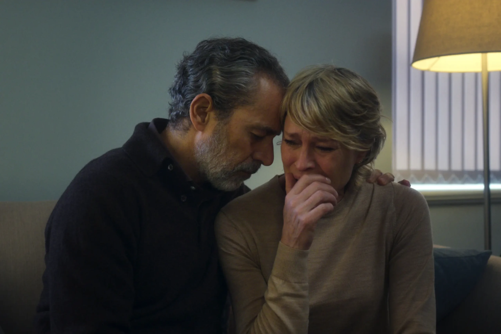 Cena da primeira temporada de The Girlfriend. Laura (Robin Wright) é consolada por seu marido, Howard (Waleed Zuaiter). À direita, de frente para a câmera, Laura é uma mulher caucasiana de meia-idade, loira, de cabelos curtos, chorando, com a mão direita cobrindo sua boca. Ela usa um suéter bege de mangas longas. À esquerda, Howard é um homem de pele escura e cabelos escuros com mechas grisalhas e uma barba grisalha, usando um suéter escuro por cima de uma camiseta cinza. Ele apoia sua testa na cabeça de sua mulher. Os dois estão sentados num sofá marrom e, atrás deles, vemos uma parede azul clara com uma janela à direita, fechada com uma cortina e iluminada por um abajur aceso.