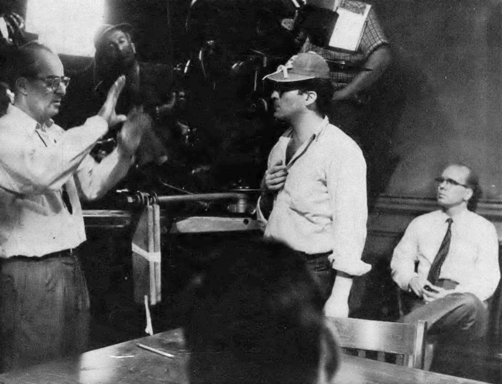 Cena dos bastidores do filme 12 Homens e uma Sentença. Em preto e branco: à esquerda, o cinemtógrafo Boris Kaufman está com as mãos levantadas, explicando algo para Sidney Lumet, à sua direita, entre a mesa que divide a sala do filme. À direita de Lumet, sentado contra a parede, está o ator John Fiedler e, atrás de Kaufman podemos ver alguns homens operando o maquinário responsável por iluminar a cena. Kaufman é um homem caucasiano de óculos e cabelos escuros, começando a rarear, usando uma camisa branca e calças claras. Lumet é um homem caucasiano de cabelos pretos, usando um boné com alguns adereços, uma camisa branca com o decote aberto e calças escuras. Ele possuí cabelos escuros e usa óculos . Fiedler, caracterizado como seu personagem, é um homem caucasiano jovem de cabelos curtos, usando óculos e vestindo uma camisa branca, com uma gravata longa e calças claras.