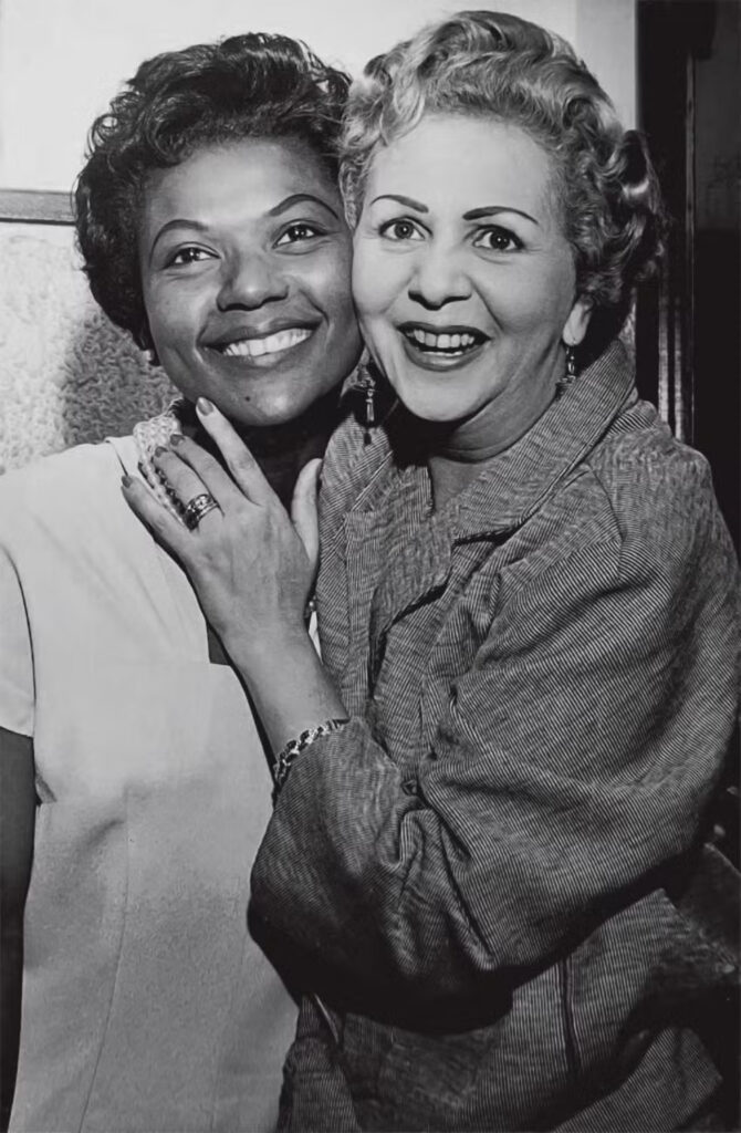 Fotografia das cantoras Alaíde Costa e Dalva de Oliveira. Imagem em preto e branco. Alaíde Costa está no lado esquerdo da imagem. Ela é uma mulher negra, olha para frente e sorri. Dalva de Oliveira está no lado direito da imagem. Ela é uma mulher parda, olha para frente e sorri.