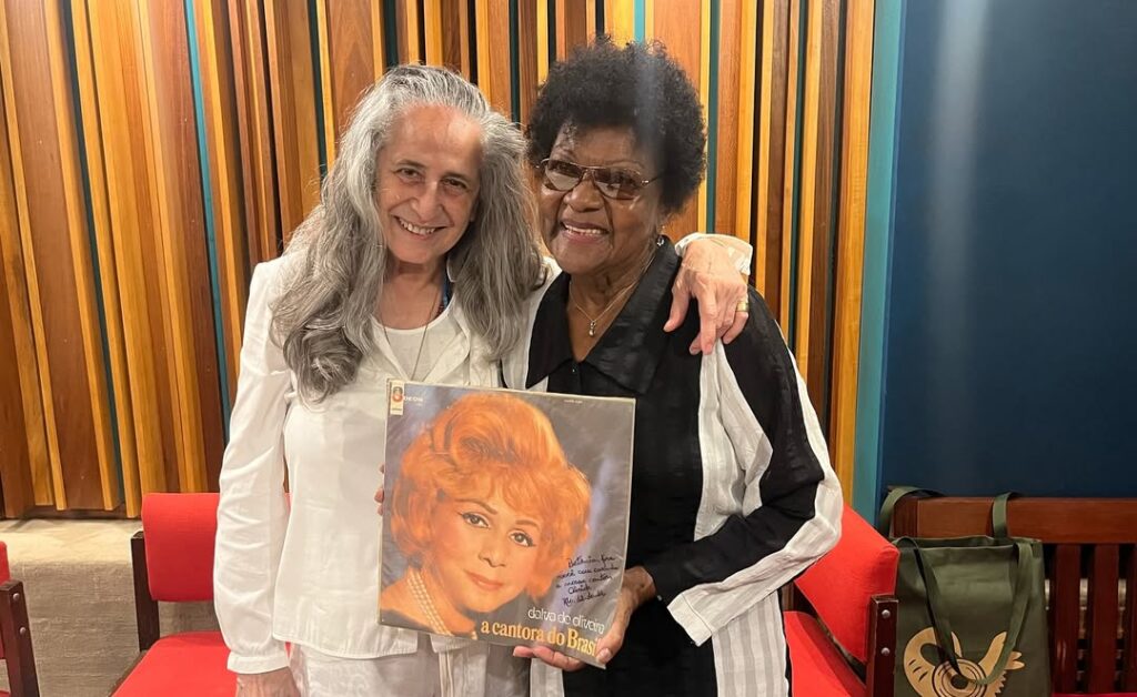Fotografia das cantoras Maria Bethânia e Alaíde Costa. Maria Bethânia está no lado esquerdo e Alaíde Costa está no lado direito da foto. As artistas olham para frente e sorriem. Alaíde segura o disco A Cantora do Brasil, de Dalva de Oliveira. O disco exibe uma dedicatória de Alaíde para Bethânia. 