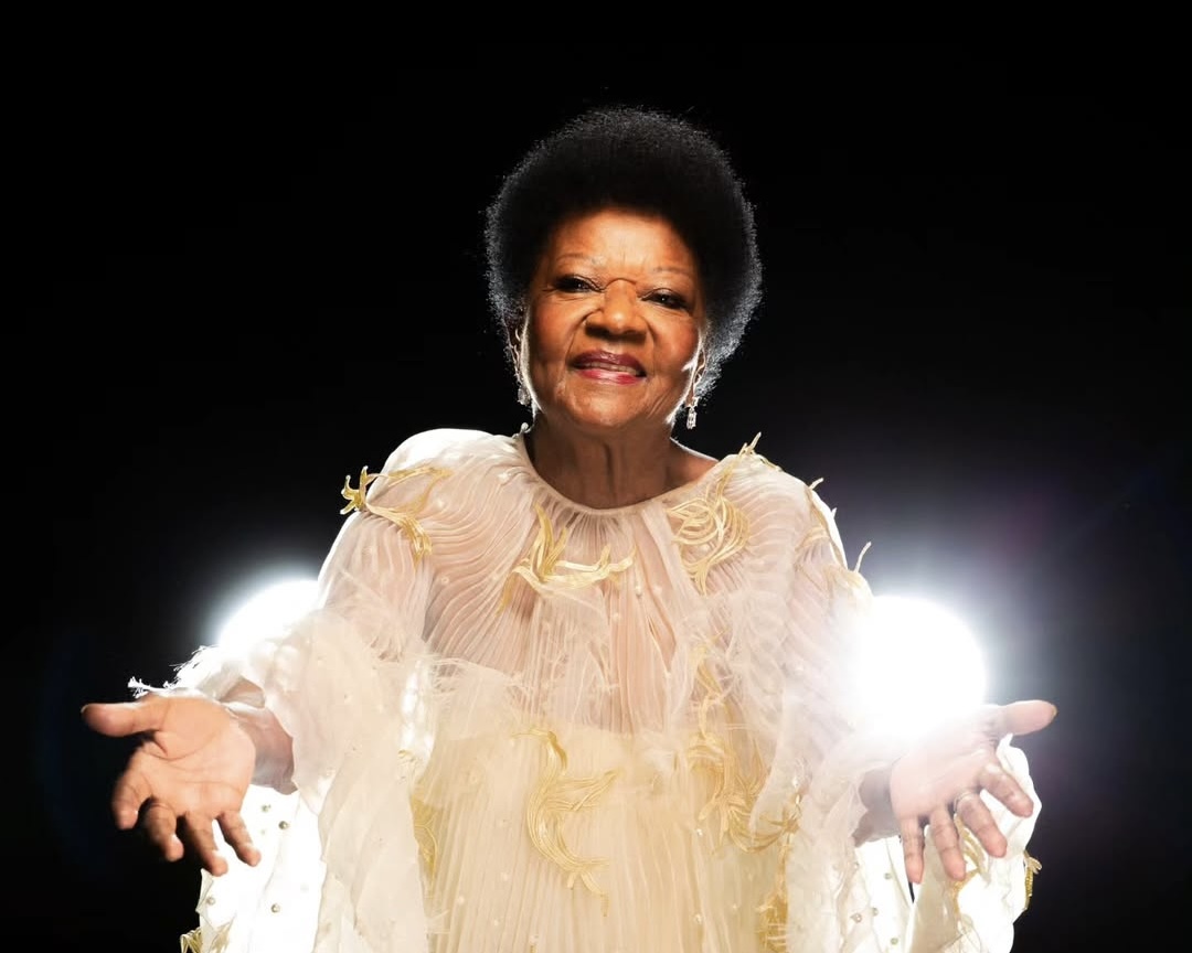Fotografia da cantora Alaíde Costa. A imagem tem um fundo preto. Alaíde aparece sorrindo, de braços abertos, usando um vestido claro. Sobre as mãos da artista, é possível ver uma forte luz branca.