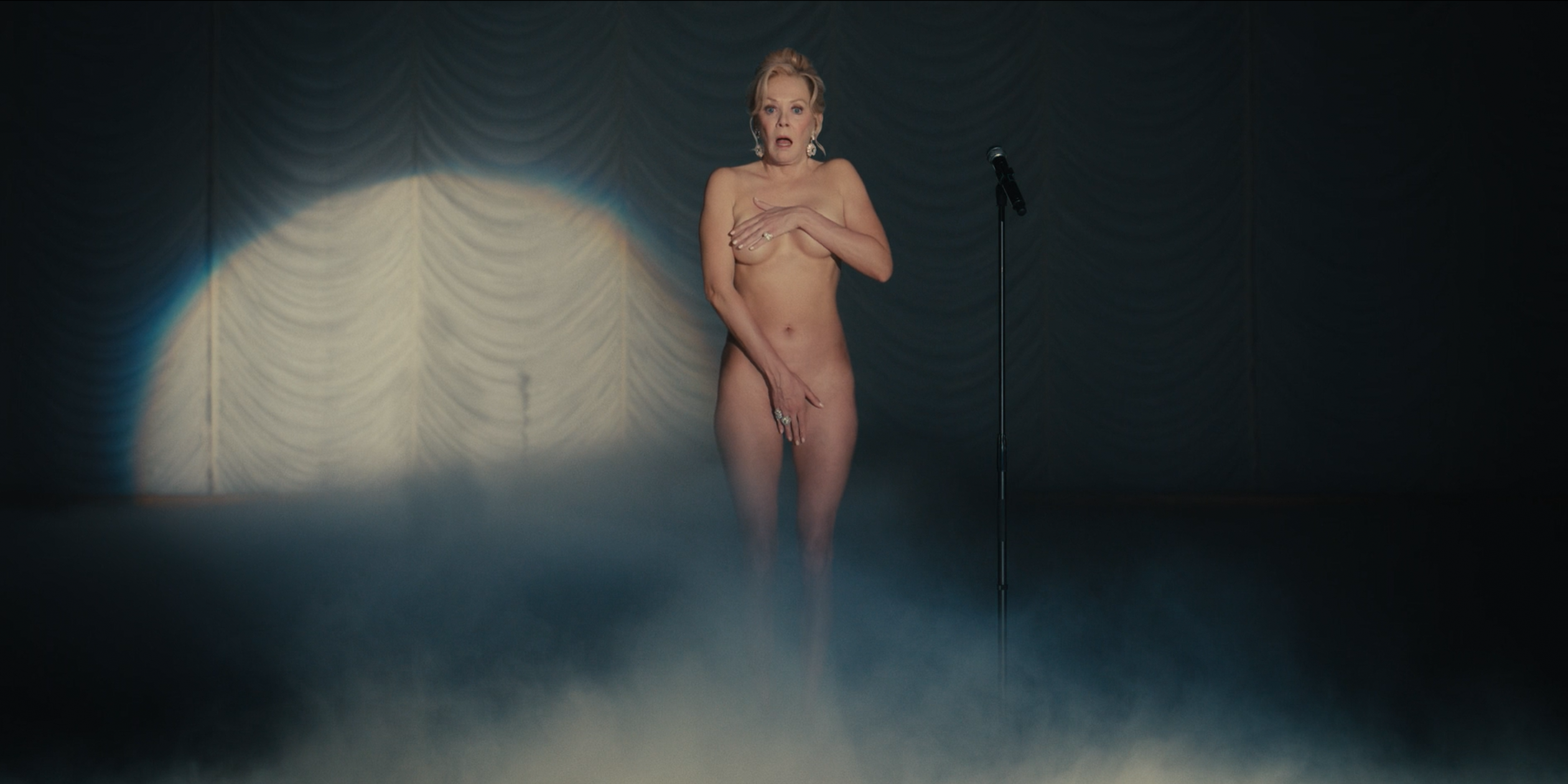 Uma mulher está sozinha no palco de um teatro, iluminada por um foco de luz diante de uma cortina escura. Ela é Deborah Vance, que cobre o peito e a região íntima com as mãos, enquanto exibe uma expressão de surpresa ou constrangimento. Ao lado, há um microfone em pedestal, e uma névoa suave cobre parte do chão, reforçando o clima dramático da cena.