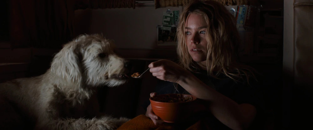 Cena do filme Supergirl. Kara (Milly Alcock), alimenta o cachorro Krypto com cereal. Kara é uma mulher caucasiana, de cabelos loiros e longos, deitada em uma cama enquanto observa algo fora da tela, levantando distraidamente uma colher de cereal com a mão esquerda e dando de comer à Krypto, seu cachorro de pelos brancos e descabelados. Atrás deles, vemos uma parede clara com algumas fotos presas.