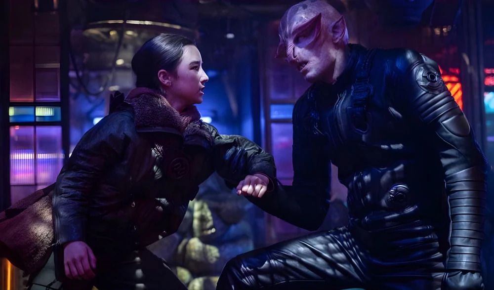 Cena do filme Supergirl. Ruthye (Eve Ridley) é agarrada por um alienígena em uma taverna. Ruthye é uma garota asiática de cabelos escuros, presos em uma trança, usando uma jaqueta escura com pelagem clara no colarinho, carregando a bainha de uma espada nas costas. Seu braço esquerdo é agarrado por um alienígena careca de pele clara, com um nariz achatado, orelhas longas e grandes protuberâncias em suas sobrancelhas. Ele usa um uniforme escuro com alças e fivelas. Atrás deles, vemos o fundo escuro da taverna, iluminado por algumas luzes azuis e roxas, painéis de vidro e outros corpos alienígenas fora de foco.