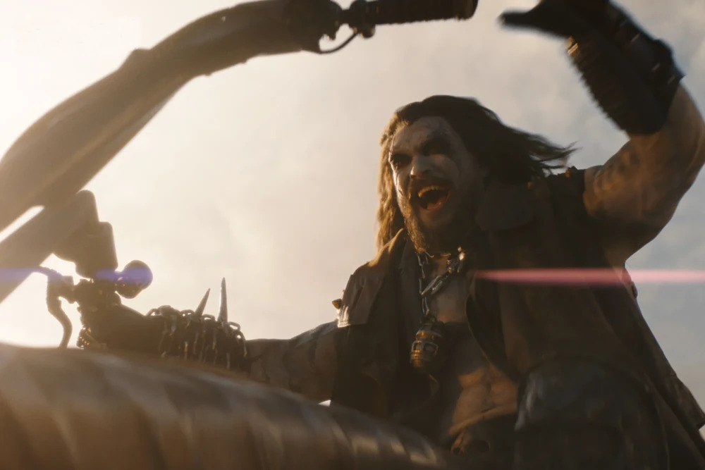 Cena do filme Supergirl. Lobo (Jason Momoa) ri em cima de sua motocicleta. Lobo é um homem de pele branca com maquiagem escura ao redor dos olhos e cabelos escuros e longos, assim como sua barba. Ele usa um colete escuro aberto, deixando seu peito visível. No pescoço, ele usa uma corrente com um objeto cilíndrico de metal na ponta. Suas mãos estão cobertas por luvas escuras, com uma corrente metálica coberta de espinhos ao redor de seu braço esquerdo.