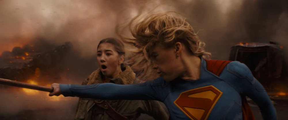 Cena do filme Supergirl. Kara (Milly Alcock) salva Ruthye (Eve Ridley) de um projétil, segurando-o com sua mão direita antes que ele a acerte. Kara, à direita, é uma mulher caucasiana de cabelos loiros e longos, usando um uniforme azul com uma insígnia vermelha e amarela na forma de um “S” no peito, com uma capa vermelha nas costas. Atrás dela, à esquerda, Ruthye é uma garota asiática de cabelos negros, presos em uma trança, usando uma jaqueta escura com pelagem clara no pescoço. Atrás delas vemos um monte de poeira levantada e alguns destroços de veículos em chamas.