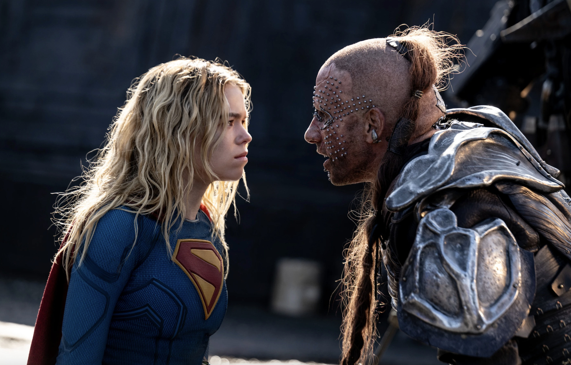 Cena do filme Supergirl. Kara (Milly Alcock) encara o bandido Krem (Matthias Schoenaerts). À esquerda, Kara é uma mulher caucasiana, com cabelos loiros longos, usando um uniforme azul com uma insígnia vermelha e amarela no peito, no formato de um “S”, com uma capa vermelha nas costas. Krem, à direita, é um homem caucasiano com a cabeça completamente raspada, à exceção de uma longa trança ruiva que vai da nuca até o torso, passando pelo lado esquerdo do pescoço. Ele usa uma armadura com pedaços de metal por cima de couro escuro e em seu rosto há pequenos pinos prateados redondos, assim como outros adereços decorando seus olhos e suas orelhas. Atrás deles, fora de foco, vemos um cenário escuro apesar do Sol que os ilumina.