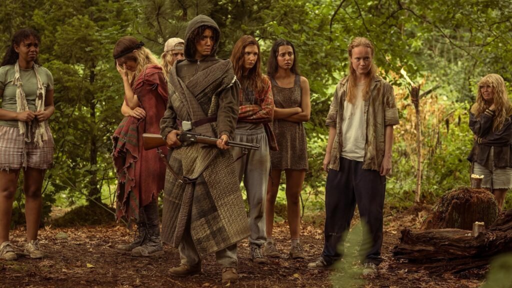 Texto alternativo: Cena da terceira temporada de Yellowjackets. As adolescentes se reúnem na floresta para planejar uma execução. Tai (Jasmin Savoy Brown) segura um rifle com as mãos, usando um edredom cinzento como túnica por cima de um moletom escuro. Tai é uma garota negra de cabelos encaracolados, cobertos pelo capuz. Ao seu lado, sua namorada, Van (Liv Hewson) olha para o seu prisioneiro, à direita. Van é uma garota caucasiana de cabelos ruivos longos, com uma cicatriz grande na face esquerda, usando uma camisa sem mangas suja por cima de uma camiseta branca e calças escuras. Atrás delas, podemos ver Natalie (Sophie Thatcher), Shauna (Sophie Nélisse) e Melissa (Jenna Burgess). Shauna e Melissa usam roupas normais e sujas, olhando severamente para a direita. Natalie olha para o lado oposto, com a mão esquerda cobrindo seu rosto, usando uma túnica de tecido vermelho. Outras garotas se reúnem ali perto, olhando com atenção. Atrás, no canto direito e distante de todas, Misty (Samantha Hanratty) olha para elas com lágrimas nos olhos.