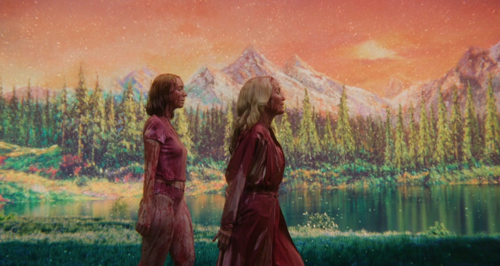 Duas mulheres com roupas manchadas de sangue caminham à margem de um lago, diante de uma floresta de coníferas e montanhas nevadas, sob um céu alaranjado e luminoso.