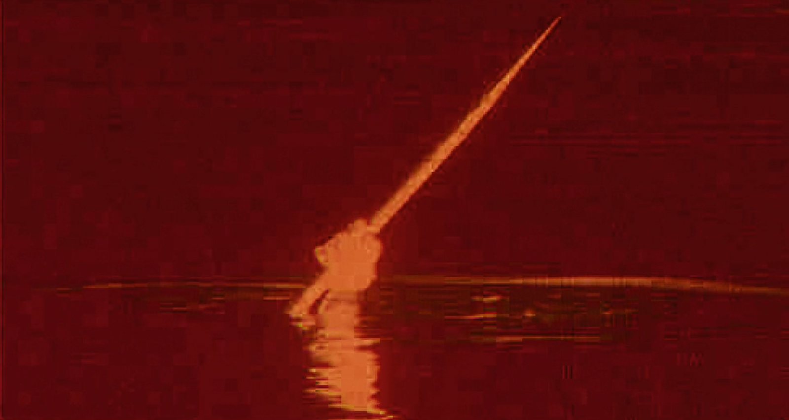 Imagem em tons de vermelho e laranja, com a silhueta de uma pessoa segurando um objeto longo e pontiagudo erguido diagonalmente. A cena apresenta distorções visuais e interferências horizontais, criando um efeito de imagem fragmentada.