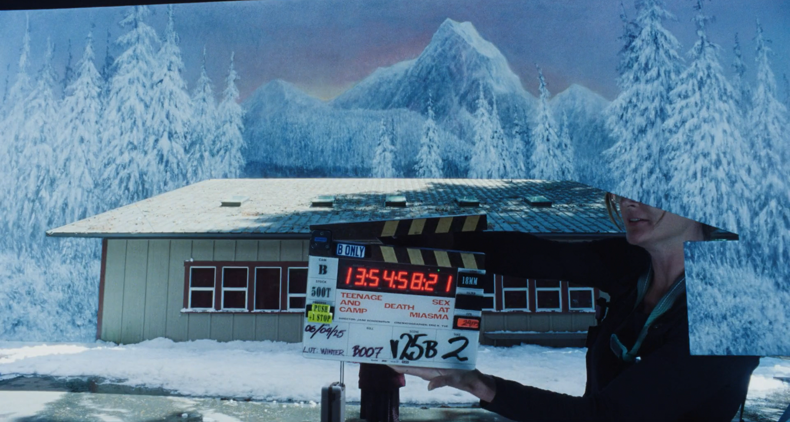Pessoa segura uma claquete de filmagem diante de um prédio baixo em um cenário coberto de neve, cercado por árvores nevadas e montanhas ao fundo. A imagem apresenta tons azulados e mostra parte do rosto e do corpo da pessoa à direita.
