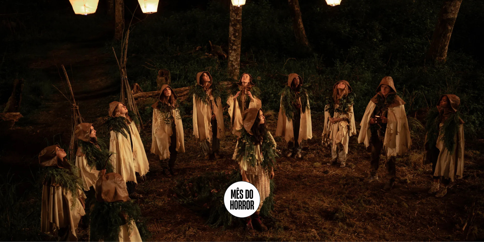 Cena da terceira temporada de Yellowjackets. Um grupo de adolescentes está reunido em um semi-círculo, dentro da clareira de uma floresta, à noite. Elas usam tecidos brancos sobre o corpo e capuzes marrons. Há apenas garotas, com exceção de Travis (Kevin Alves), quase ao fim da linha, à direita. No centro da formação, Lottie (Courtney Eaton) olha para o alto assim como suas colegas. Ao redor delas, vemos estruturas tribais como fogueiras, cercados e bancos e, ao fundo, o resto da floresta escura.