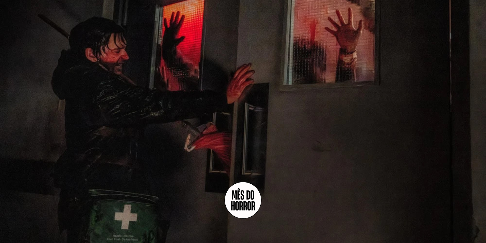Cena do filme Resident Evil. Bryan (Austin Abrams) bloqueia uma porta contra uma investida de zumbis. Bryan é um jovem caucasiano de cabelos escuros e roupas de inverno escuras, coberto de sangue, colocando as mãos contra as portas cinzas de um hospital. Ele carrega uma bolsa verde nos ombros com uma cruz branca. Através das janelas nas portas, vemos mãos se chocando contra o vidro e vultos disformes. O sangue manchado nas janelas dá uma luz vermelha à cena.