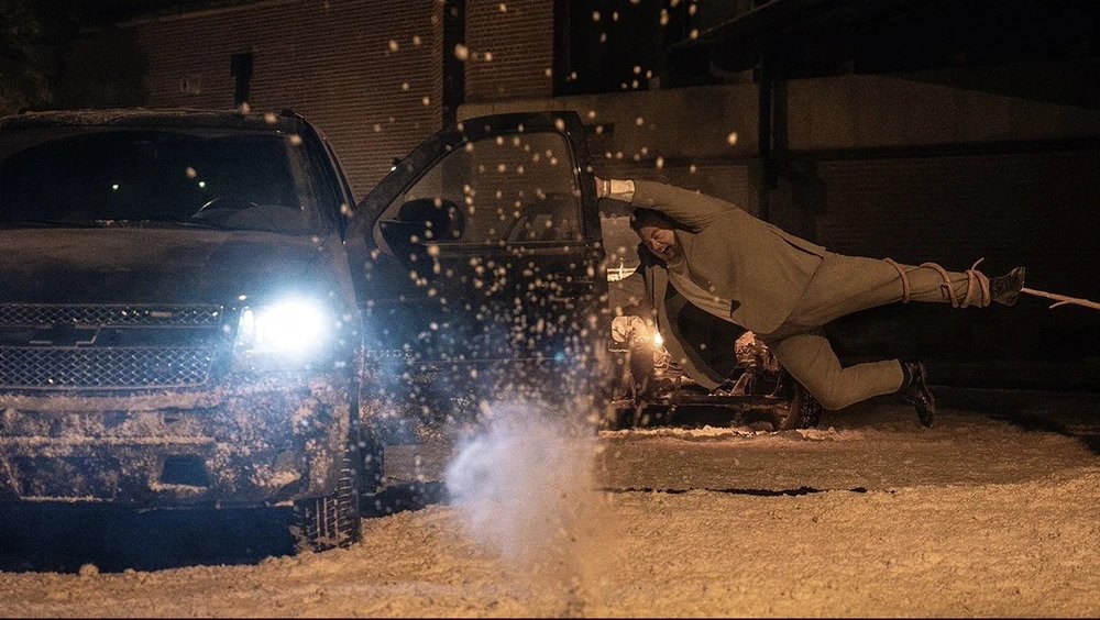 Cena do filme Resident Evil. Paul (Paul Walter Hauser) é arrastado para fora de seu carro por um tentáculo vindo da direita. Paul é um homem caucasiano e pesado, usando um terno cinzento. Ele se segura na porta aberta do motorista, enquanto o tentáculo puxa sua perna esquerda. Os faróis de seu carro ainda estão ligados; é um carro preto e grande coberto por uma fina camada de neve. O chão está coberto por neve e, atrás deles, vemos uma parede de tijolos iluminada pela luz amarela de postes.