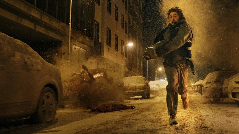 Cena do filme Resident Evil. Bryan (Austin Abrams) corre numa rua nevada conforme corpos caem dos céus nos carros estacionados. Bryan é um jovem caucasiano de cabelos escuros, usando vários casacos e abraçando uma bolsa térmica verde. A cena é iluminada pela luz amarela vinda dos postes de iluminação.